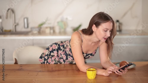 Woman is standing near table in the kitchen, she is looking at the marketplace website and choosing new clothes. Woman drinking coffee in the morning in the kitchen with a smartphone in her hands 