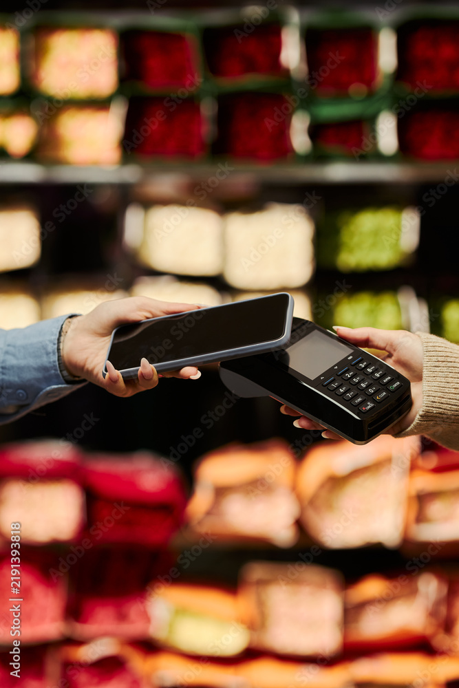 Obraz premium Vertical image of young customer paying with smartphone via contactless POS terminal at grocery store. Scene illustrating NFC digital transaction, fintech mobile banking