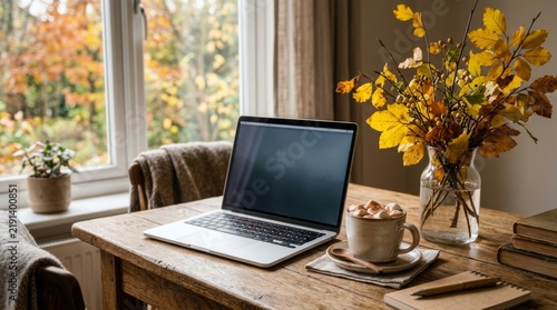 Laptop with hot cocoa and marshmallows on rustic wooden table near window with autumn foliage. Cozy fall home office with yellow leaves in vase, blanket and books.