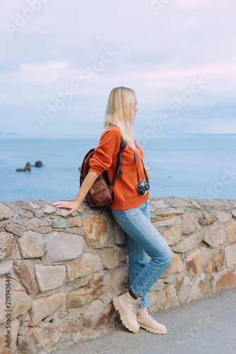 Fotografie Young woman traveler with backpack and vintage camera leaning on a stone wall, e