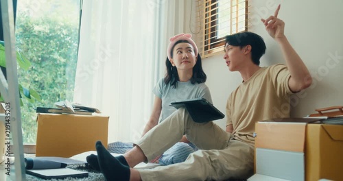 Young Asian couple plans move at home while browsing furniture online on tablet beside packed boxes. Relocation planning, new home setup, online shopping, household design and unpacking concept.
