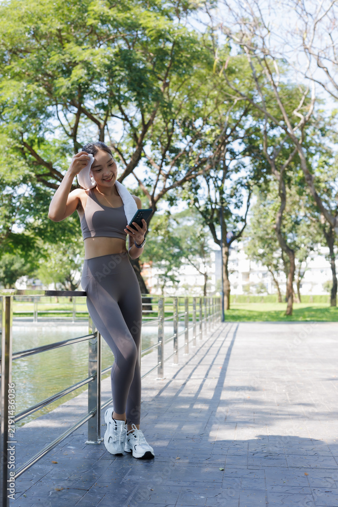 Fototapeta premium Young Asian woman pausing during her workout in a green park, wiping sweat from her forehead with a towel and engaging with her smartphone for fitness tracking