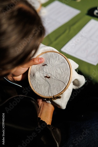 Close-up of a person holding an embroidery hoop with light fabric, dark thread, and small insect motifs during a hands-on outdoor craft activity