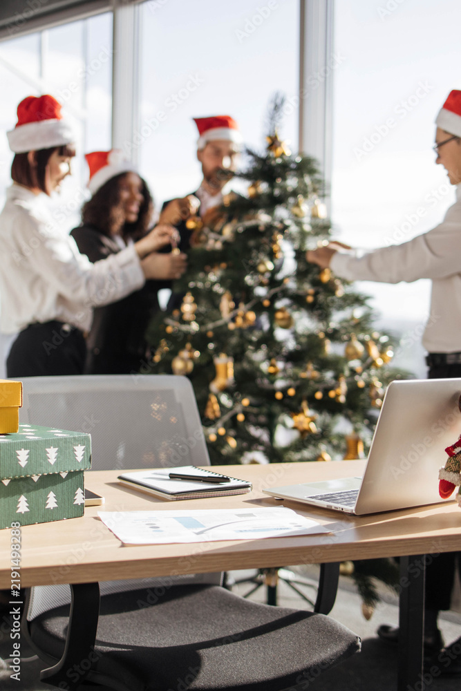 Fototapeta premium Colleagues in Santa hats decorate a Christmas tree in a modern office setting with gifts and a laptop on a desk
