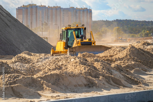 Quadro su tela Heavy yellow bulldozer leveling sand mound on a road construction site with dust in the air and high-rise residential building in background