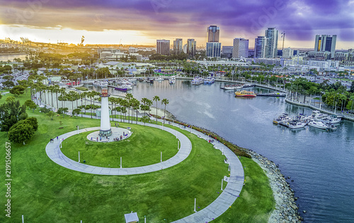 Aerial view of Lion Lighthouse for Sight in Shoreline Aquatic Park at Rainbow Harbor, with marina boats, green lawns, and city skyline under a sunset sky in Long Beach, CA, United States.