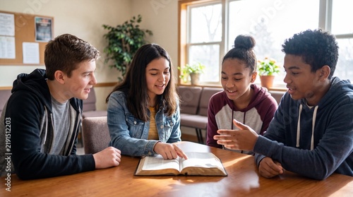 Diverse group of teenagers reading book together at table. Young man and women studying bible in sunny room with plants and window. Faith, friendship and youth education concept.