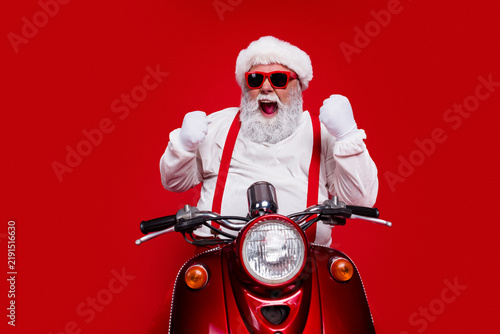 Excited Santa in red sunglasses and suspenders riding a vintage red scooter for Christmas celebration and holiday fun