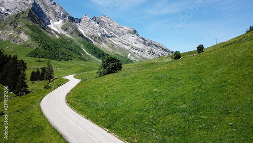 Aerial view of the winding road at Col des Aravis surrounded by lush green alpine meadows and dramatic mountain peaks in La Clusaz, Auvergne-Rhone-Alpes, France.
