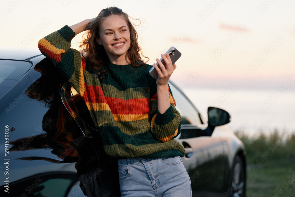 Obraz premium A smiling young woman holding a smartphone, leaning against her car by the sea during sunset. Concept for travel, modern lifestyle, and freedom on a road trip.