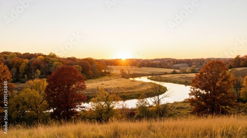 Sunset over a winding river in an autumn landscape. Sunburst on the horizon across colorful fall foliage and rolling hills. Autumnal equinox scenic countryside view