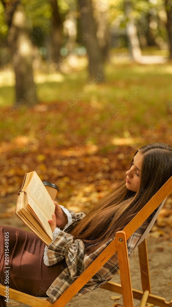 Obraz premium Young woman relaxing in a chair reading a book in a park during autumn