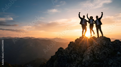 Silhouettes of diverse team members celebrating success on mountain peak at sunset with arms raised