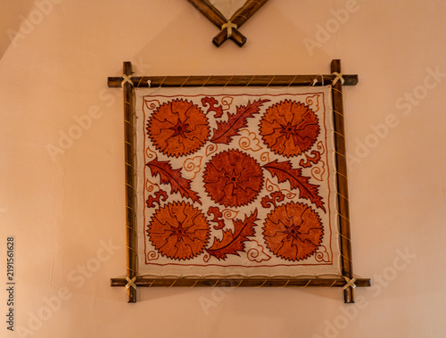 A traditional Uzbek embroidered textile featuring vivid orange floral and leaf motifs is displayed on a wooden frame against an indoor wall, highlighting Central