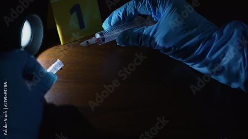 Evidence collection, a gloved hand capping a bloody syringe and taking it away from a wooden floor with dried blood next to a yellow evidence marker under a side flashlight and police lights.