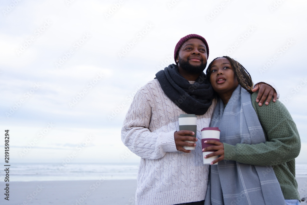 Obraz premium Couple standing close on beach holding cups, wearing knit sweaters, scarves and beanie, copy space