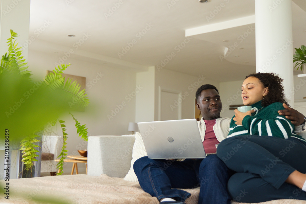 Obraz premium African American couple sitting on sofa in living room, holding laptop, cradling mug, copy space