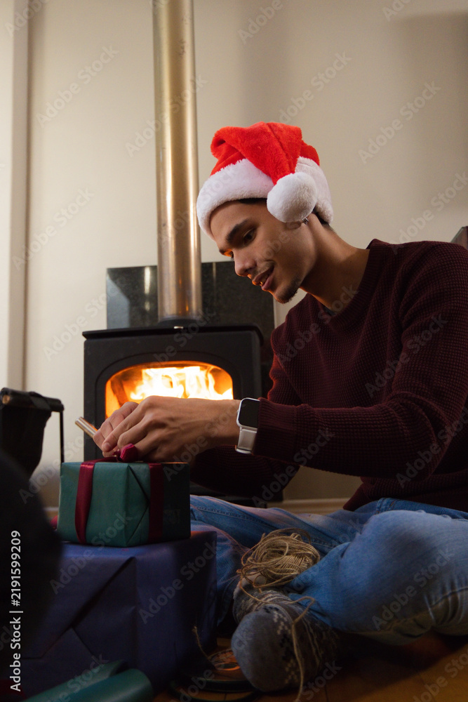 Obraz premium African American man wearing red Santa-hat sitting on hardwood floor tying ribbon on gift by stove