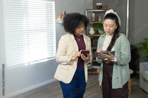 Diverse female coworkers in blazer reviewing on phone and tablet with stylus near window