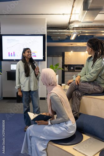 Diverse coworkers in biz casual presenting, taking notes in office with mic and charts on screen