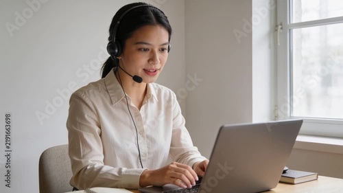 Typing woman in blouse joining call at desk near window, speaking into headset and adjusting laptop