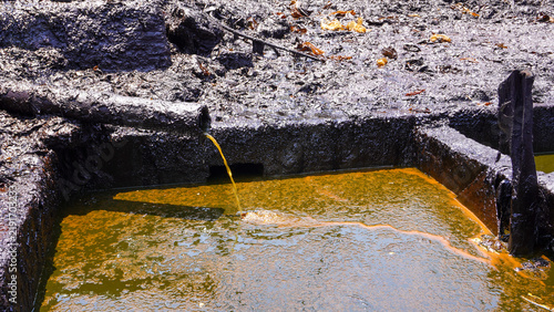 Raw crude oil pouring from a pipe into a reservoir pool at traditional mining site in Wonocolo, Bojonegoro, Indonesia. Shows energy production, fossil fuel extraction, and industrial impact.
