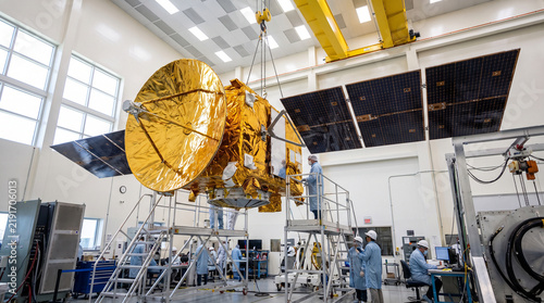 Large communications satellite covered in gold thermal foil with large gold antenna dish and deployed solar panels suspended by overhead crane in bright aerospace clean room factory