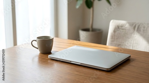 Closed silver laptop and a coffee cup on a wooden table in a bright, minimalist home office interior