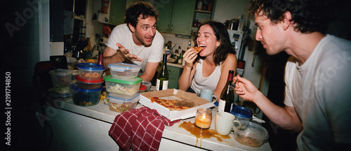 Three young adult friends hanging out in a messy kitchen at night. Group eating pizza and drinking wine in a cluttered apartment. Authentic candid lifestyle snapshot of roommates laughing and talking
