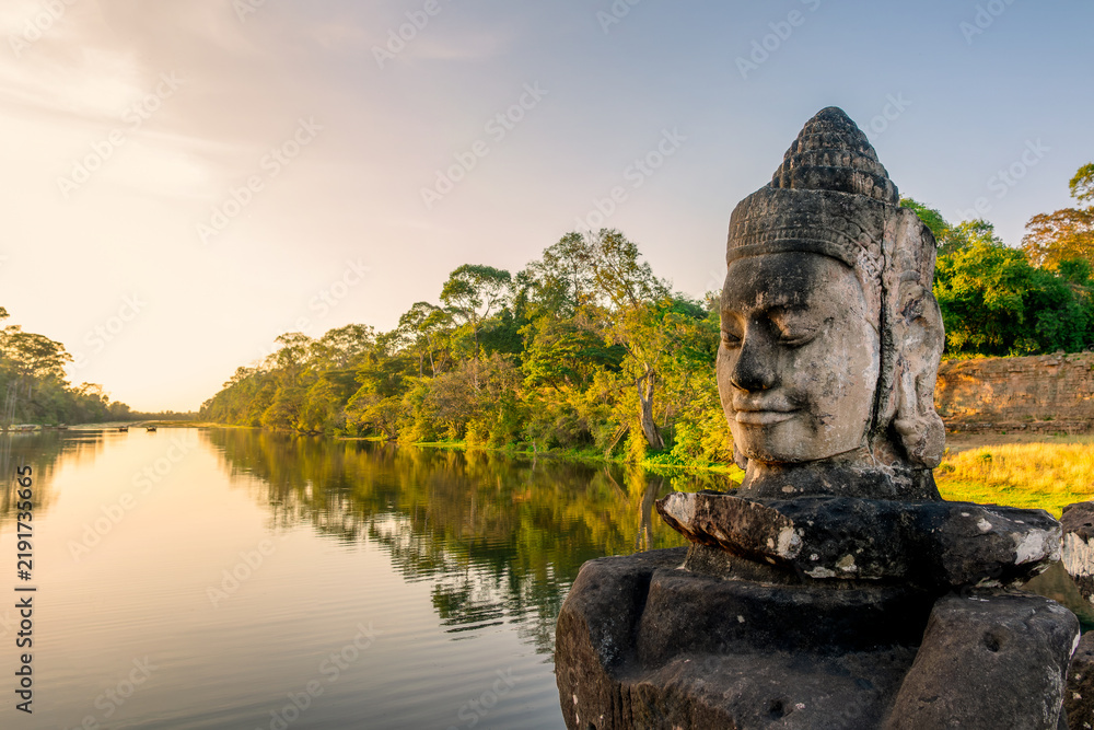Obraz premium Deva head statue at Angkor Thom South gate, smiling serene Buddha face at sunset in Angkor, ancient moat with water reflections, Siem Reap, Cambodia