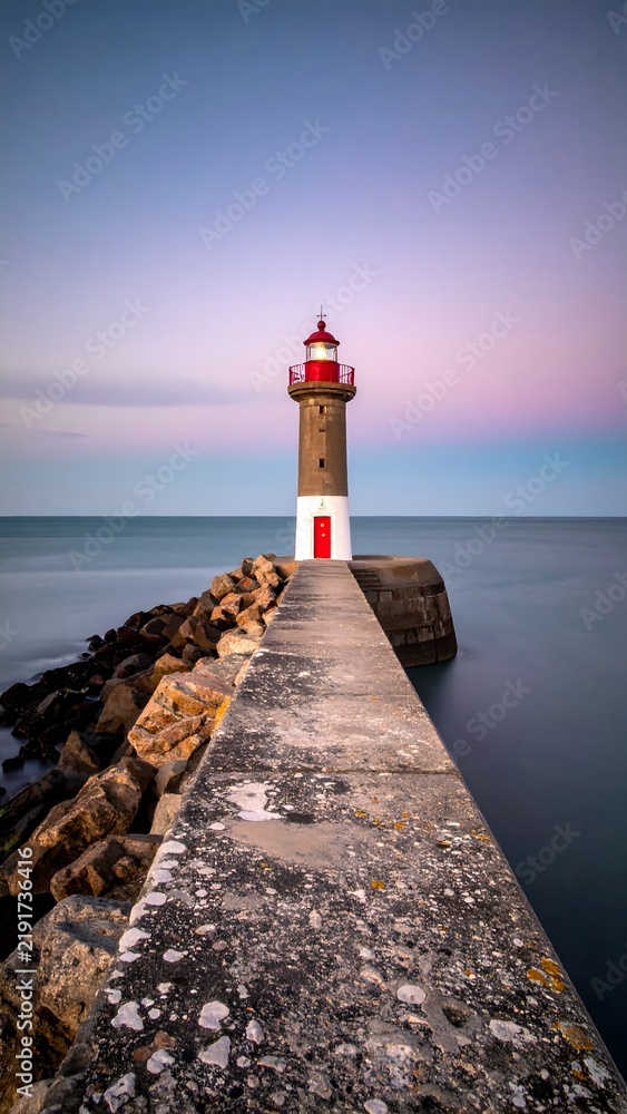 Fototapeta premium Tall Coastal Lighthouse on Rocky Pier with Calm Sea and Sky at Dusk