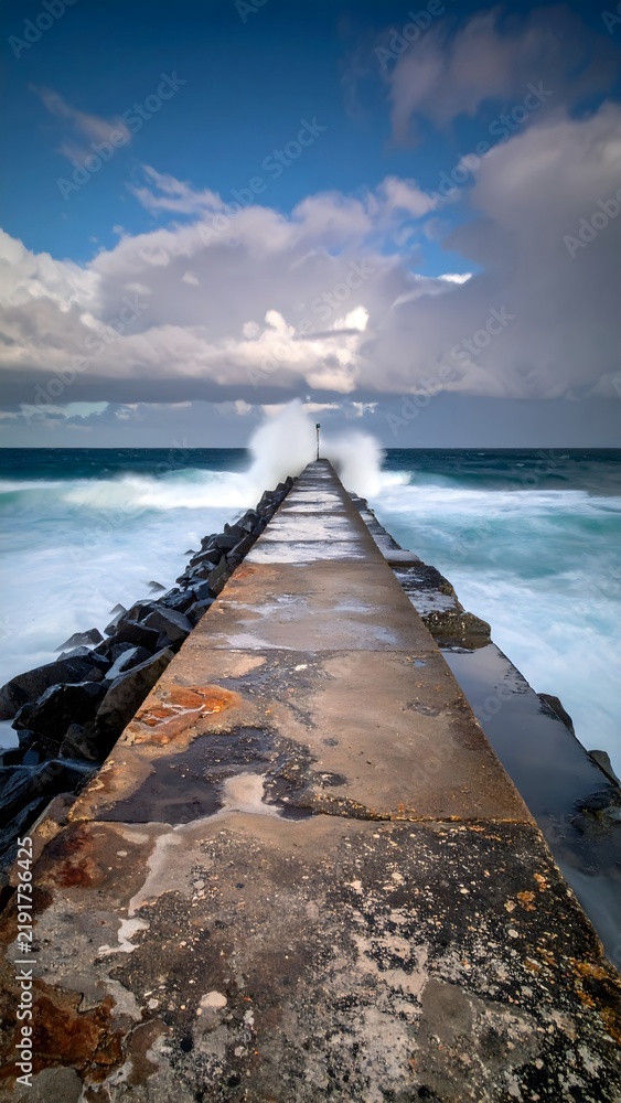 Fototapeta premium Long Pier Extending Into Ocean With Waves Splashing Against It Under Cloudy Sky