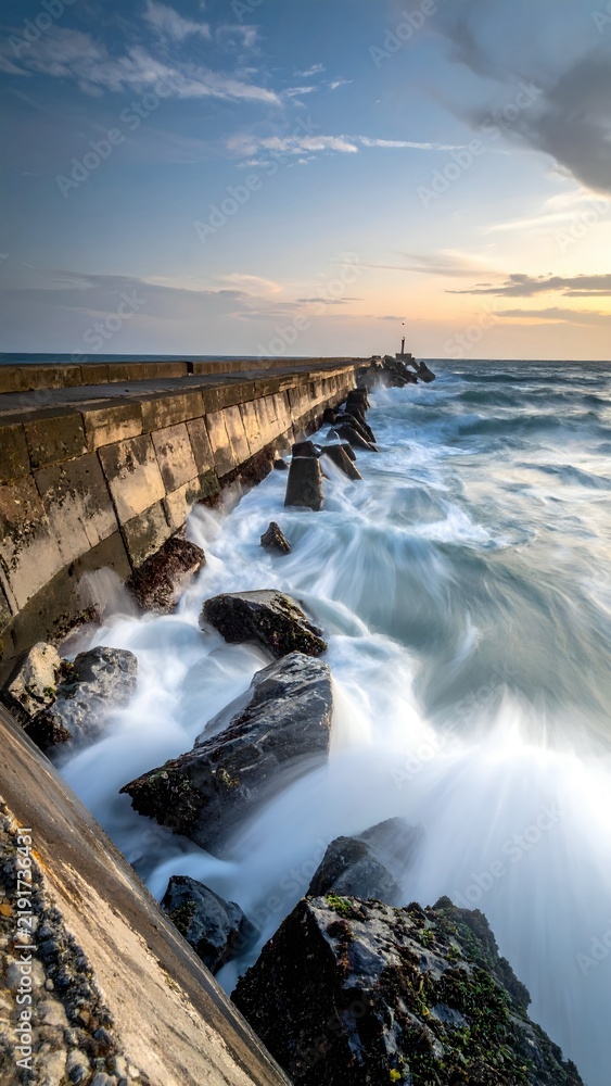 Fototapeta premium Coastal Sea Wall with Waves Crashing Against Rocks at Sunset