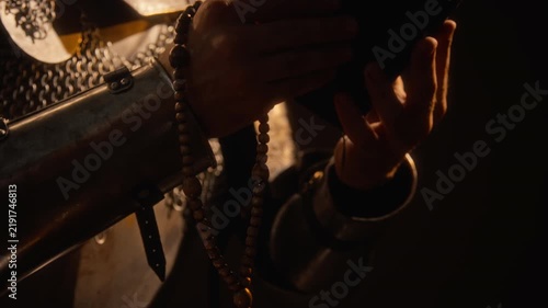 Tilt up shot of young adult knight in medieval armor holding wooden rosary beads and holy book. Clip fits historical documentaries, cinematic trailers, fantasy game teasers, and educational videos