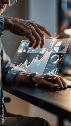 Man interacts with a transparent digital interface displaying glowing white line graphs and circular charts. Soft bokeh accents the dark indoor setting while dim warmth highlights skin texture.