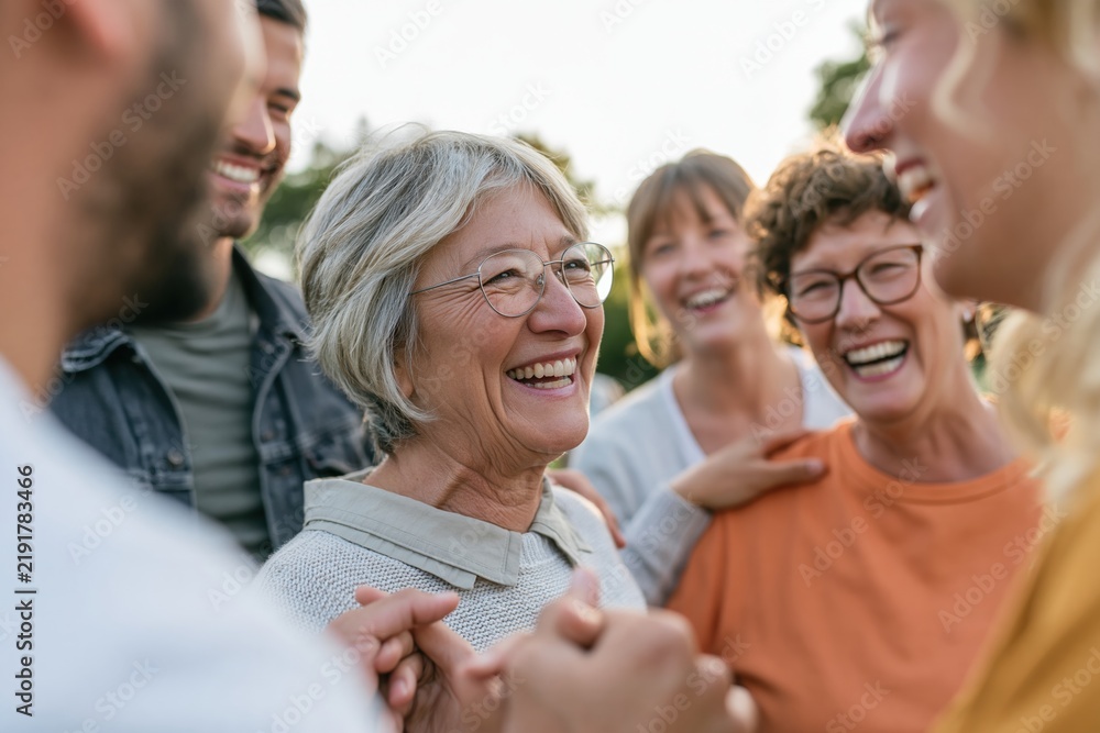 Fototapeta premium Senior woman with glasses smiling candidly among younger friends in a park, featuring soft lighting for community and intergenerational friendship