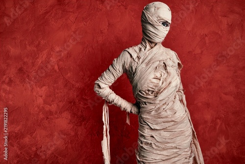 Full body portrait of mummy figure in textured bandages standing against deep red wall with one eye visible