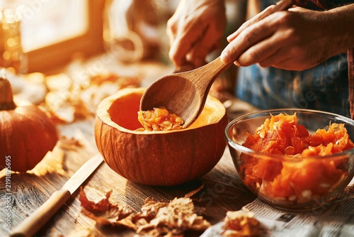Top-down close-up of hands scooping orange pumpkin seeds and pulp into glass bowl on wooden table with carving knife, hollowed pumpkin, and scattered autumn leaves during cozy DIY prep