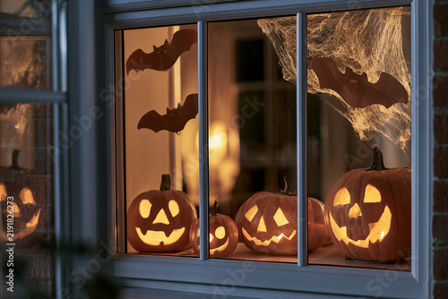 Old window night view of several carved jack-o-lantern pumpkins glowing warmly on interior windowsill with hanging paper bats and cobweb decorations