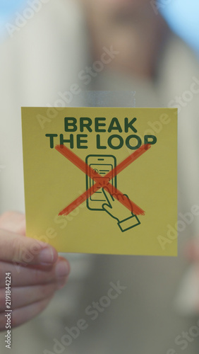 Person holding a yellow sticky note with break the loop text and a crossed out smartphone icon, symbolizing a call to reduce screen time and internet usage for well being