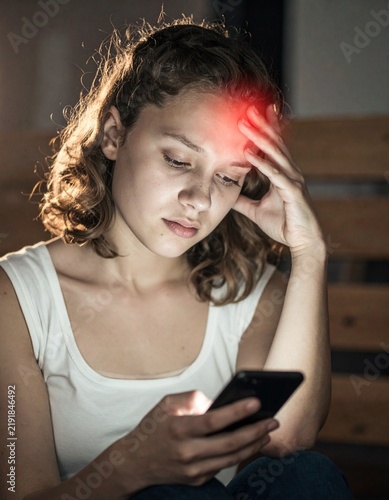 Woman experiencing a severe migraine and headache while looking at a glowing smartphone in a dark room.