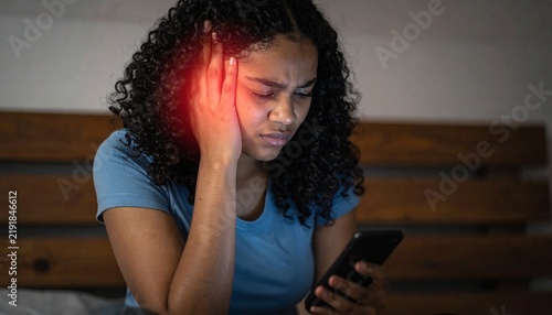 Young woman holding painful head and looking at mobile phone display with glowing light in bedroom at night.