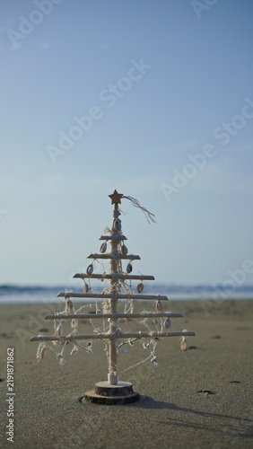 Wooden Christmas tree decorated with natural seashells and string standing on a sandy beach against a clear blue sky, symbolizing sustainable, eco-friendly, and tropical holiday celebrations
