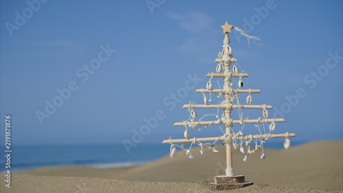 Handmade wooden Christmas tree decorated with seashells and rope standing on a sand dune, ocean waves breaking in the background under a clear blue sky, representing eco-friendly holiday travel