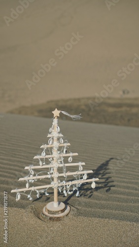 Small natural wood Christmas tree decorated with seashells standing on a ripple textured sand dune, representing holidays, travel, and eco-friendly celebrations