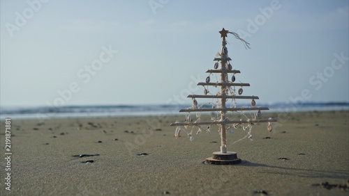 Wooden Christmas tree made of driftwood, decorated with shells and rope, standing on sunlit sand by the ocean with gentle waves and a clear blue sky in the background