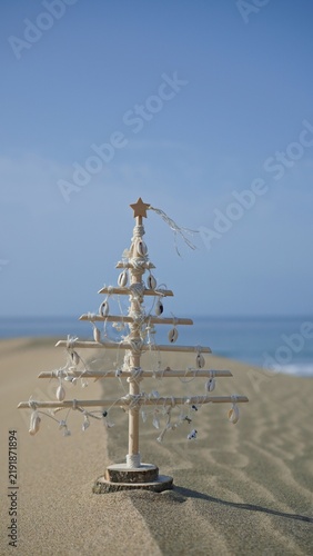Small handmade Christmas tree crafted from natural wood and seashells standing in a sand dune with the ocean in the background, representing sustainable holiday travel
