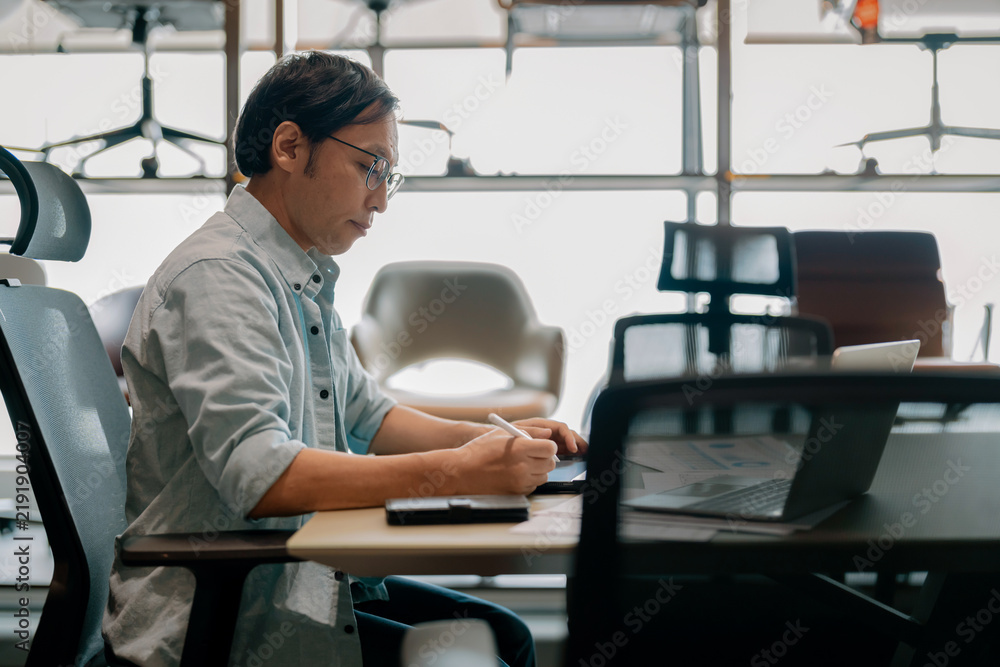 Fototapeta premium Focused Asian architect drawing on digital tablet with stylus at office desk under warm ambient light, demonstrating concentrated diligence, creative drafting precision, thoughtful planning, absort