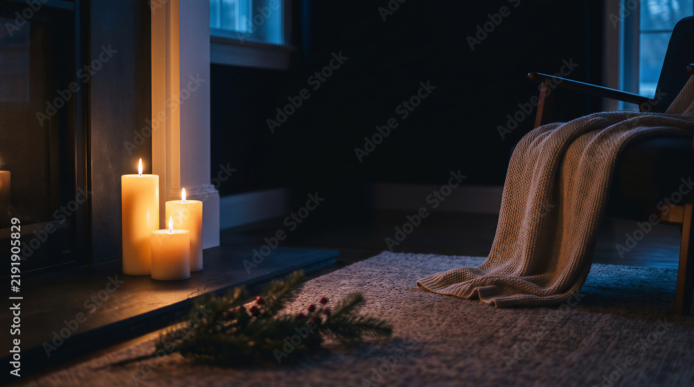 Fototapeta premium Cozy interior scene with lit candles on a fireplace hearth, a warm knit blanket on an armchair, and evergreen sprigs on a rug, creating a peaceful atmosphere with copy space