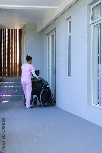 African American caregiver in pink scrubs pushing Asian senior in wheelchair at door, copy space